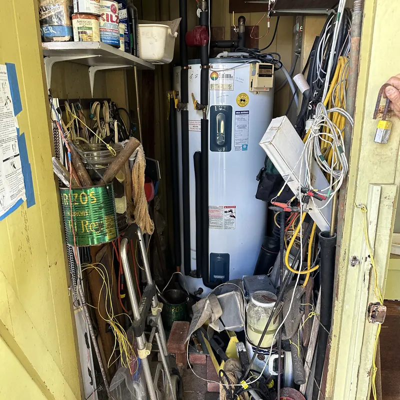 An old water heater in a cluttered utility closet alongside tools and coiled wire.